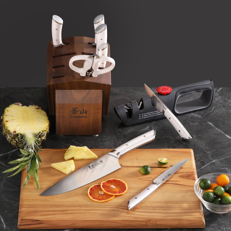 Knife block set with six knives and scissors in acacia wood block, two knives on cutting board with sliced citrus and pineapp