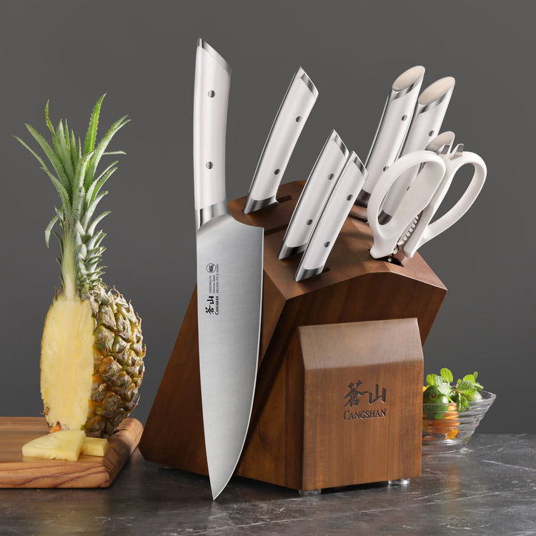 Wooden knife block holding eight stainless steel knives and kitchen scissors, next to a sliced pineapple on a cutting board.