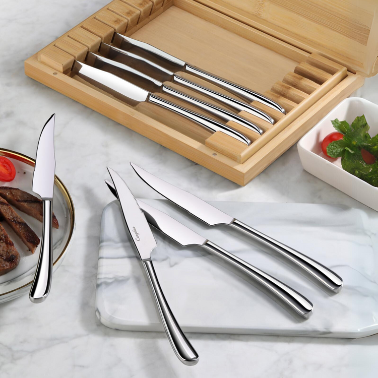 Set of eight stainless steel steak knives with curved handles, displayed in a bamboo storage box and on a marble surface.