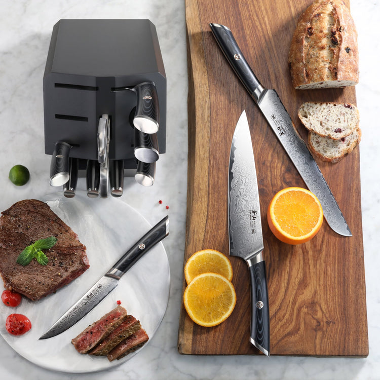 12-piece knife set with black handles in a block, displayed with sliced bread, steak, and orange on marble and wood surfaces.