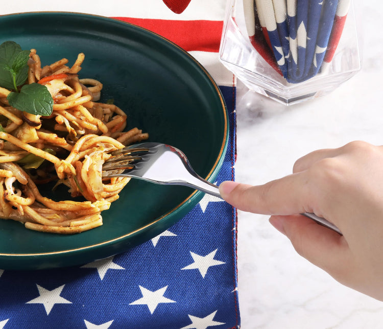 Hand holding a stainless steel dinner fork twirling spaghetti on a dark green plate over a star-patterned cloth.