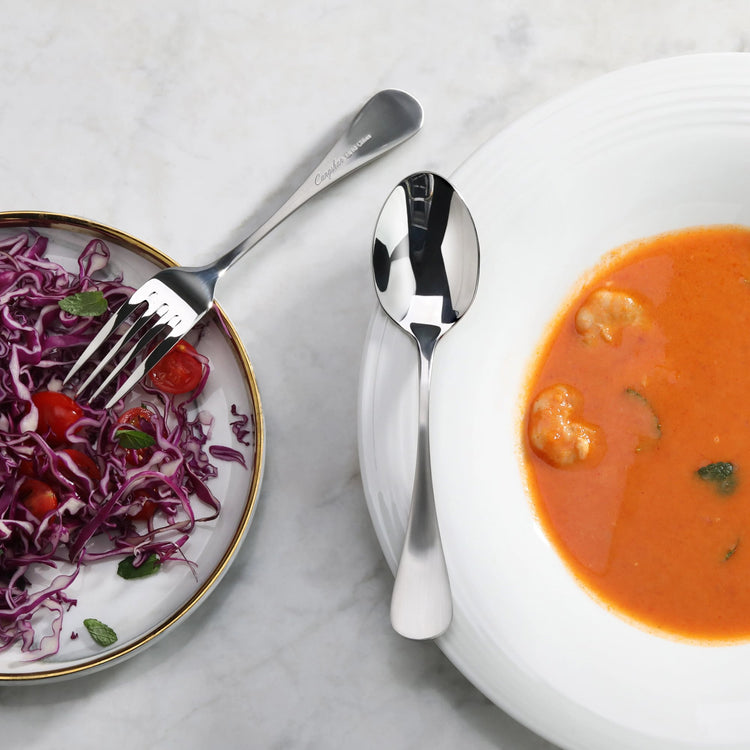 Polished stainless steel dinner spoon with matte finish handle placed on a white plate beside a bowl of orange soup and a sal