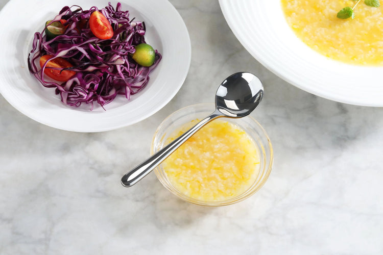 Forged stainless steel bouillon spoon resting on a small bowl of yellow soup, with salad and soup bowls nearby.