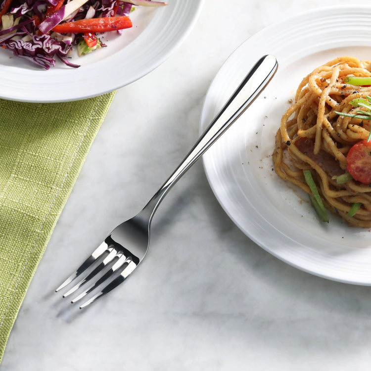 Fork with a mirror finish resting on a marble surface between plates of salad and pasta.