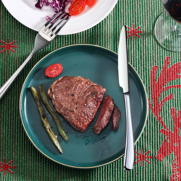 Dinner plate with cooked steak, grilled green beans, and cherry tomato slice, next to a RAIN II Series Dinner Knife with mirr