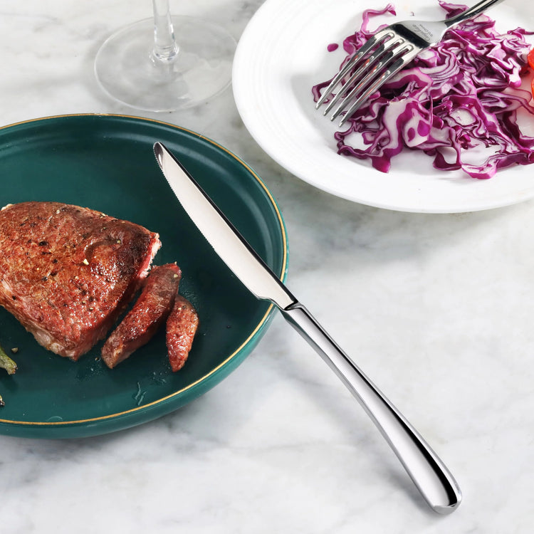 Polished stainless steel dinner knife resting on a green plate with sliced steak, next to a white plate with purple cabbage s
