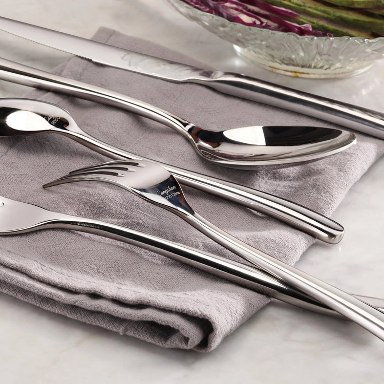 Forged stainless steel flatware set with mirror finish, including dinner spoon, fork, and knife on a gray napkin.