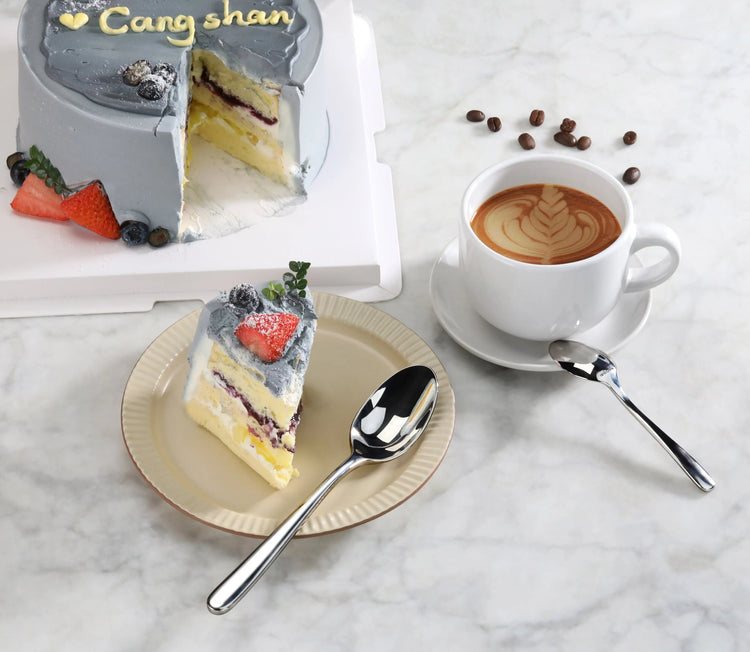 Slice of layered cake with gray frosting and berries on a plate, a cup of latte with latte art, and two stainless steel spoon