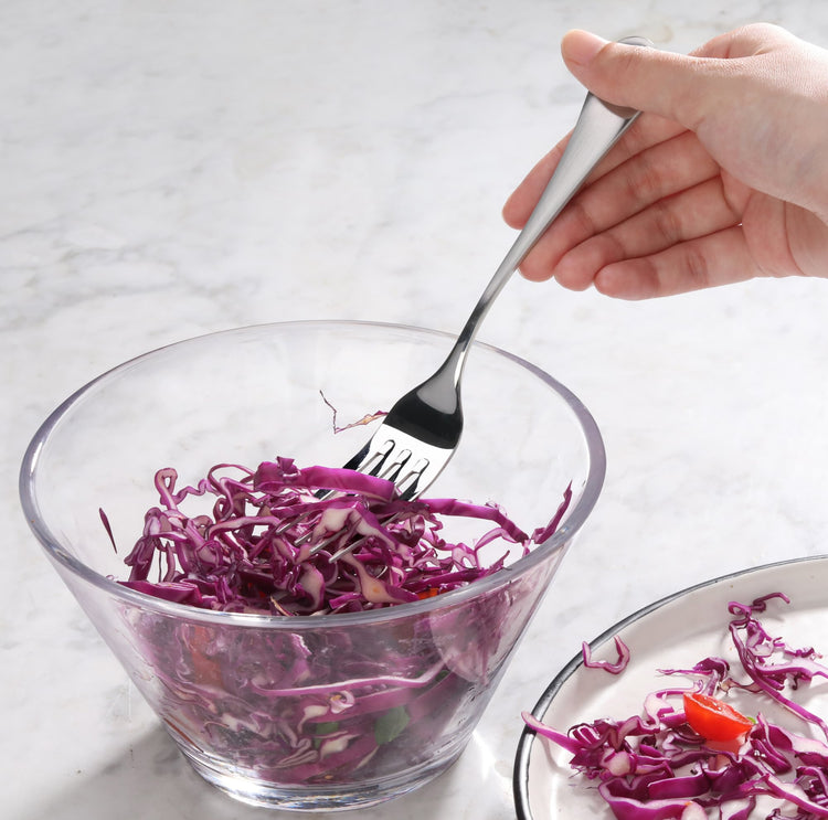 Hand holding a stainless steel salad fork with matte finish handles, mixing shredded purple cabbage in a glass bowl.