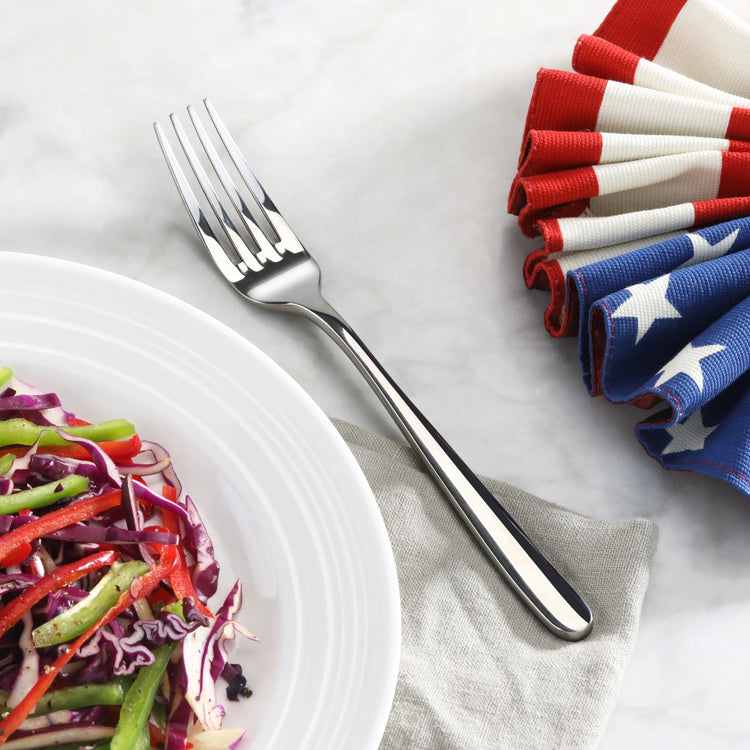 Salad fork with mirror finish resting on a gray napkin beside a plate of salad and folded American flag napkins.