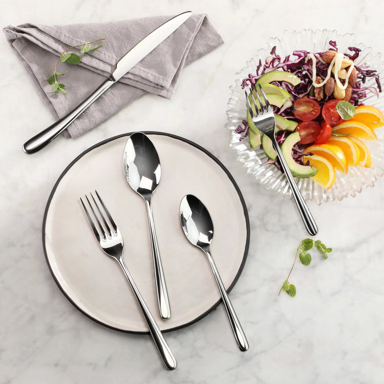 Three stainless steel utensils—a fork, a tablespoon, and a teaspoon—on a round plate, with a knife on a napkin and a salad wi