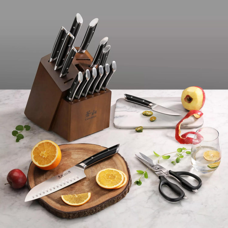 Wooden knife block with 17 knives featuring black handles, displayed with sliced oranges, apple, scissors, and a glass of wat