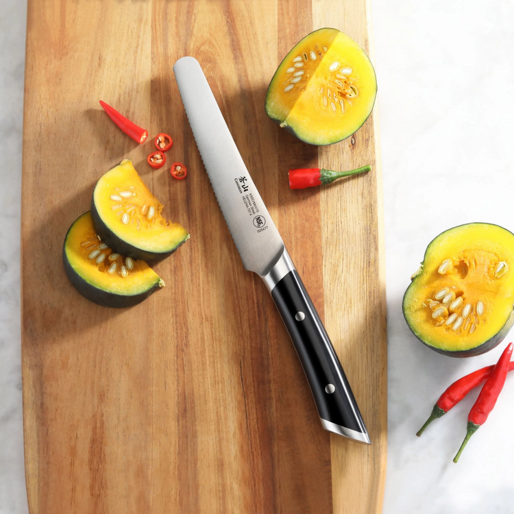 Serrated utility knife with black handle on wooden cutting board next to sliced pumpkin and red chili peppers.