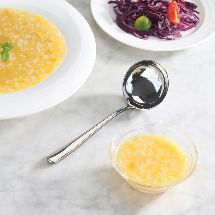 Forged stainless steel soup ladle with mirror finish resting on a marble surface near bowls of soup and salad.