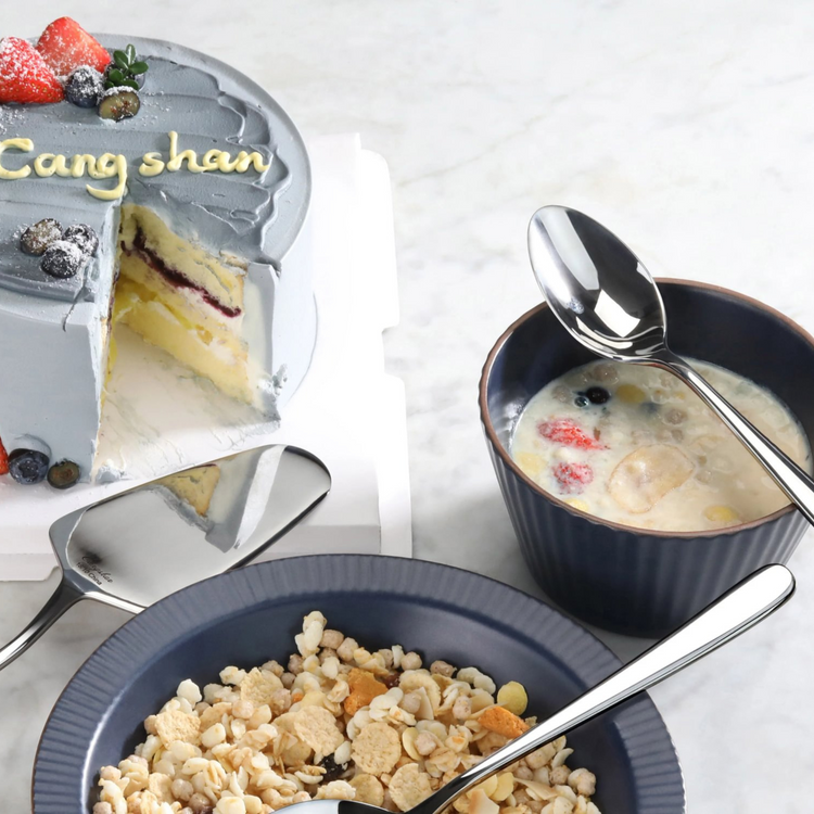 Three forged stainless steel spoons with mirror finish placed near a bowl of cereal, a cup of milk with fruit, and a sliced c