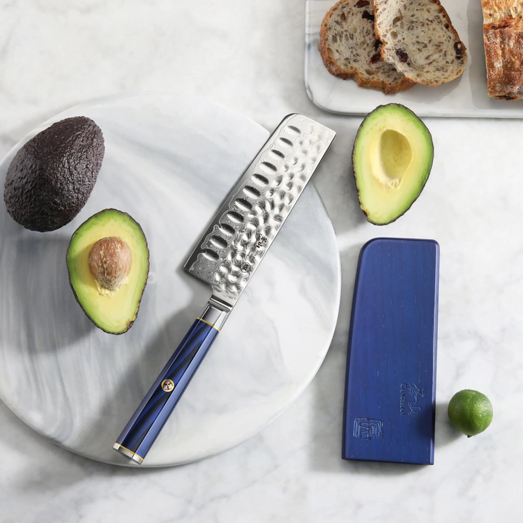 Nakiri knife with hammered blade and blue handle on marble plate, surrounded by avocado halves, bread, and lime.