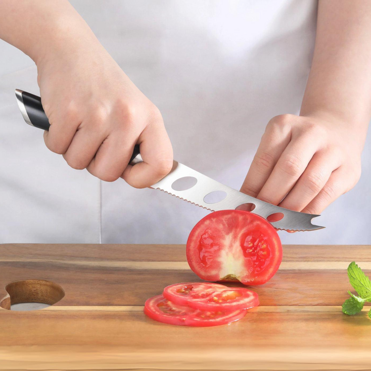 Hand holding a serrated knife slicing a tomato on a wooden cutting board with three tomato slices and a mint leaf nearby.