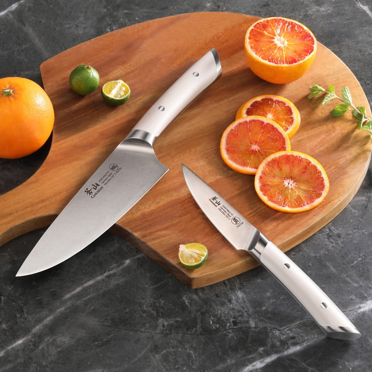 Two kitchen knives with white handles on a wooden cutting board beside sliced blood oranges.