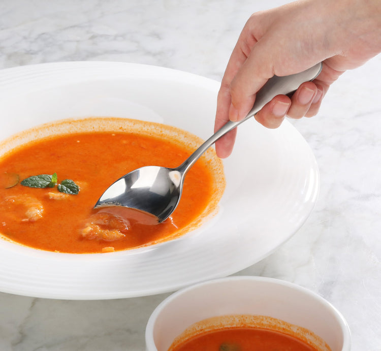 Hand holding a spoon with a matte finish handle scooping tomato soup from a white bowl on a marble surface.