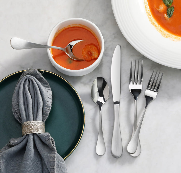 Stainless steel flatware set with knife, spoon, and two forks beside a bowl of tomato soup and a dark green plate with a gray