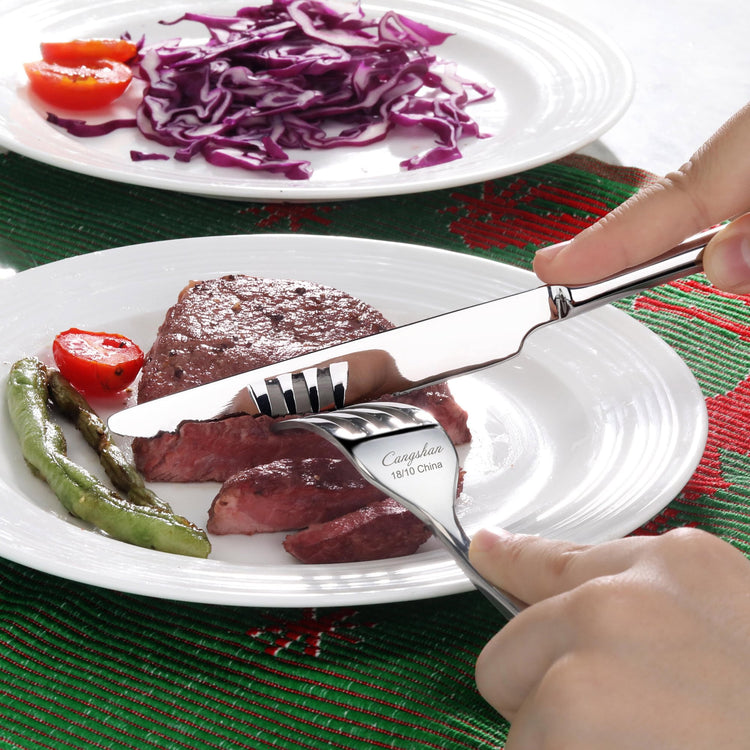 Hand holding a fork and knife cutting a cooked steak on a white plate with green pepper and cherry tomato.