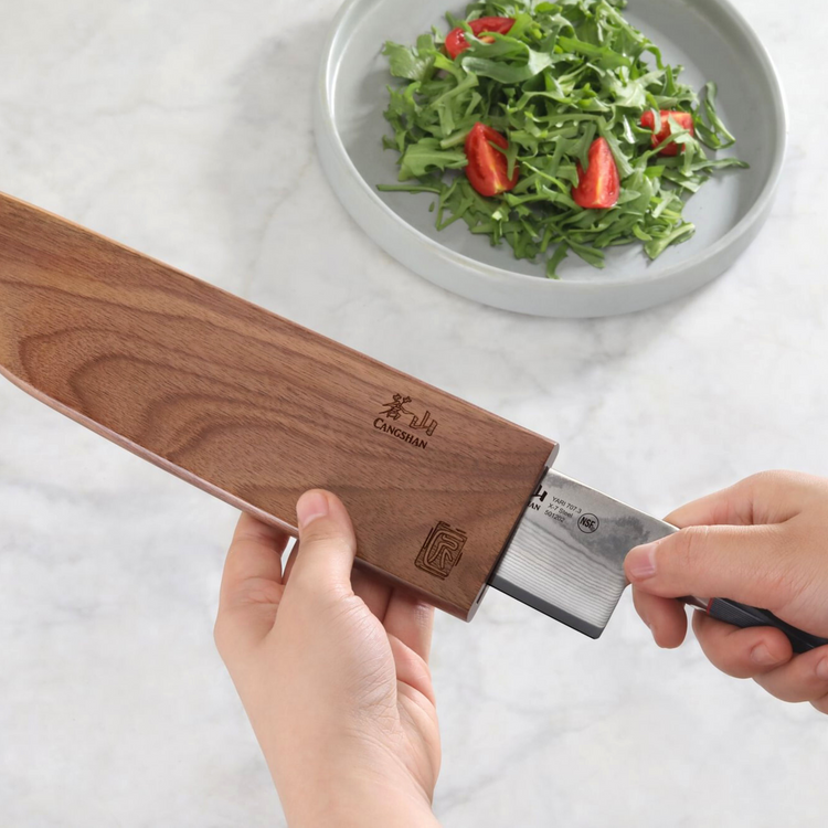 Hands sliding a knife blade into a walnut magnetic sheath with a plate of salad in the background.