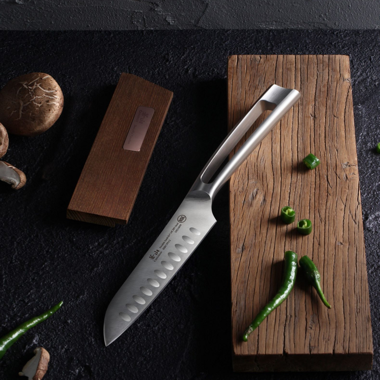 Santoku knife with hollow edge and open handle on wooden board next to ash wood sheath and green chili peppers.
