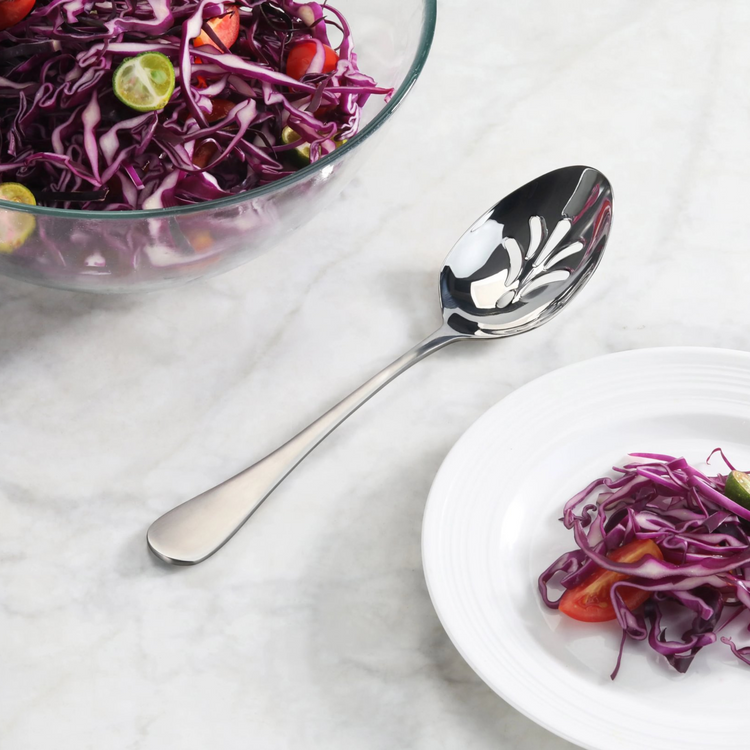Slotted stainless steel serving spoon with matte finish handle next to a bowl and plate of purple cabbage salad.