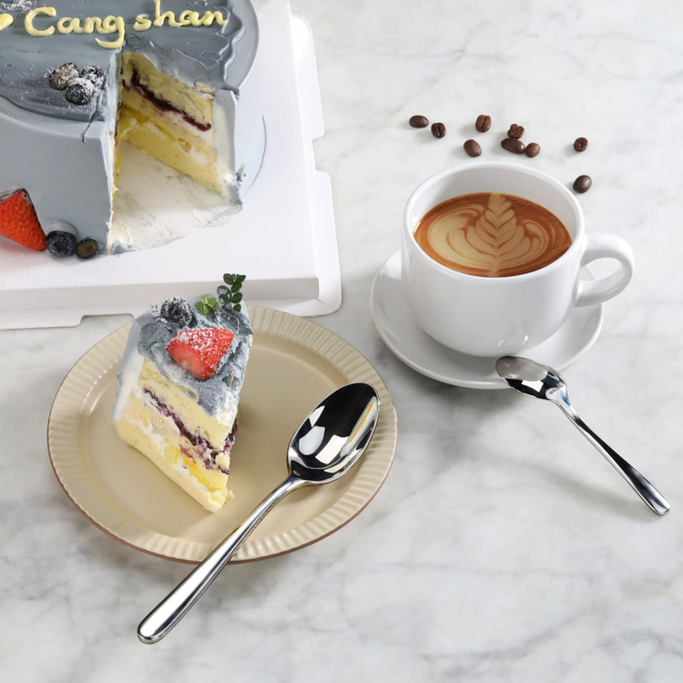 Slice of layered cake with gray frosting and berries on a plate, a teaspoon, and a cup of latte with coffee beans nearby.