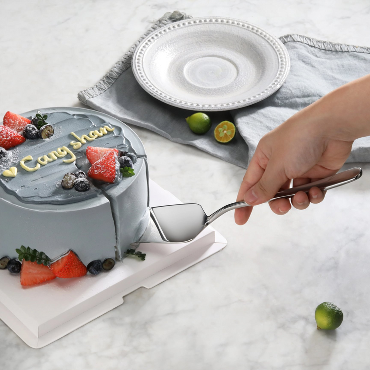 Hand holding a stainless steel pie server lifting a slice from a blue frosted cake decorated with strawberries and blueberrie