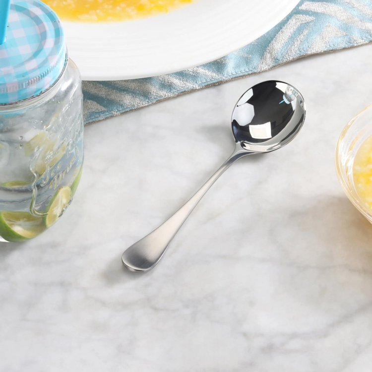 Stainless steel bouillon spoon with matte finish handle resting on a marble surface near a jar and a bowl.
