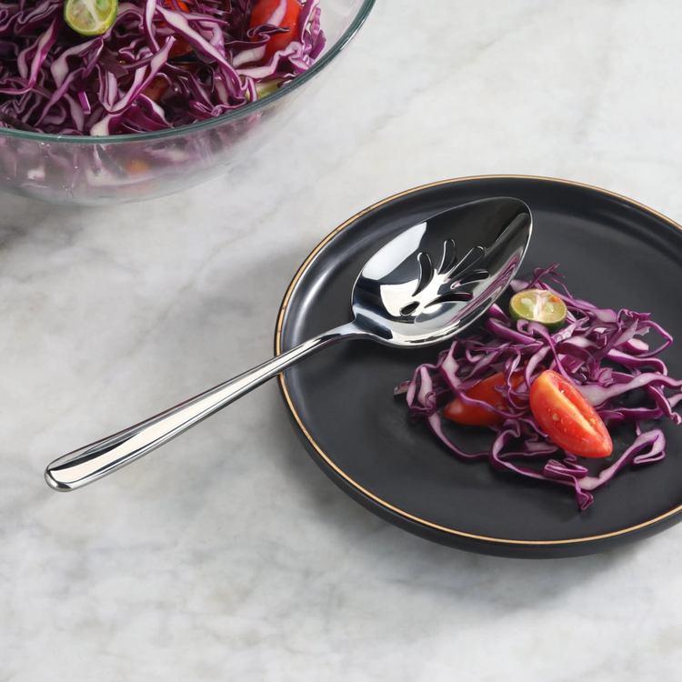 Slotted stainless steel serving spoon with mirror finish on a black plate with red cabbage and tomato slices.