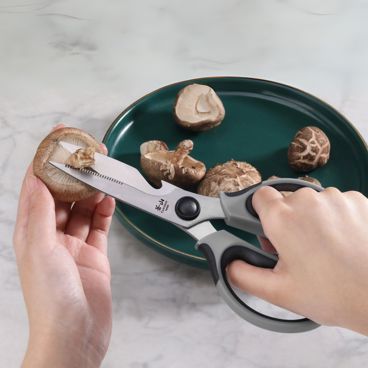 Hands using gray kitchen shears to cut the stem off a mushroom over a green plate with more mushrooms.
