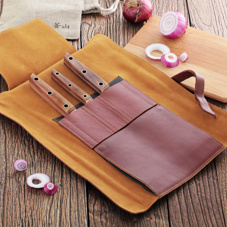 Three knives with wooden handles in a brown leather roll-up case on a wooden surface with sliced onions nearby.