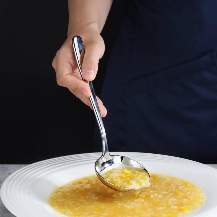 Hand holding a polished stainless steel soup ladle scooping yellow soup from a white bowl.