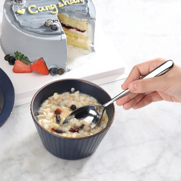 Hand holding a polished stainless steel spoon scooping oatmeal with dried fruit from a dark bowl, with a sliced cake in the b