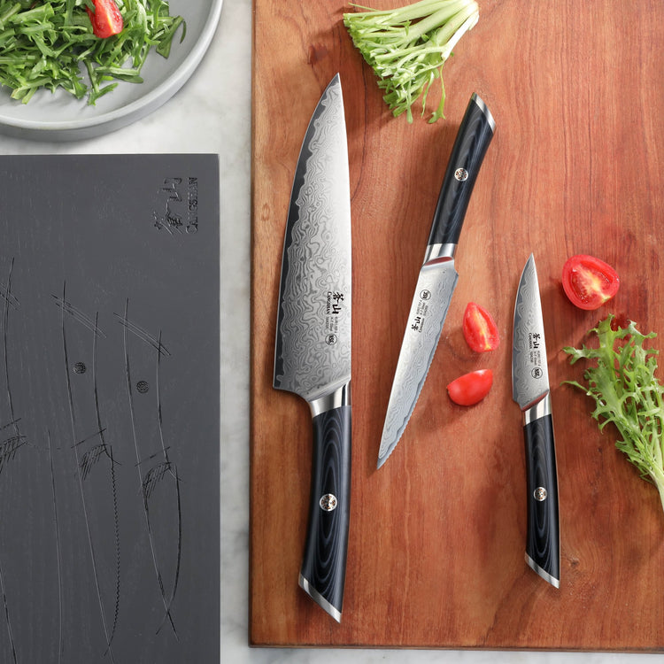 Three knives with black handles and patterned blades on a wooden cutting board with tomato pieces and greens nearby.