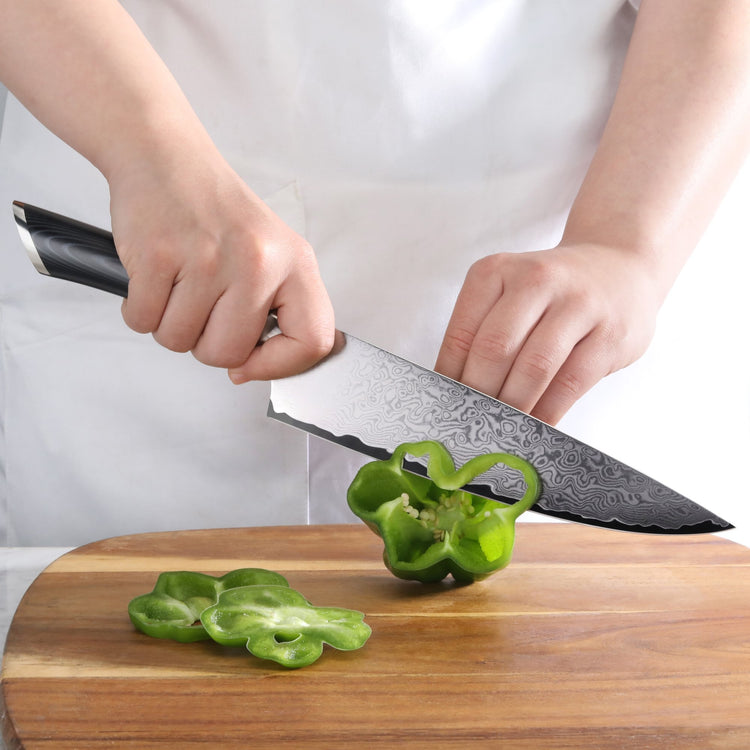 Hands slicing a green bell pepper on a wooden cutting board with a patterned Damascus steel knife.