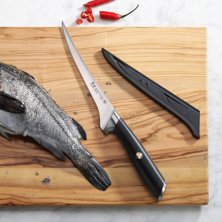 Three-piece fillet knife set with black handles and sheath on wooden cutting board next to whole fish and chili peppers.