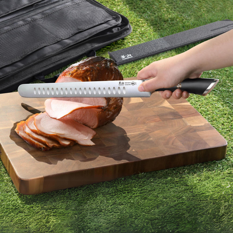 Hand holding a long serrated knife slicing cooked meat on a wooden cutting board outdoors with a black knife bag nearby.