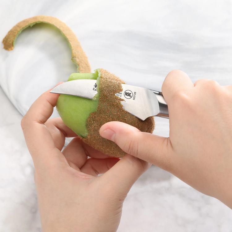 Hands peeling a kiwi fruit with a small curved stainless steel knife on a white surface.
