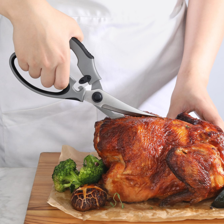 Person using gray heavy-duty kitchen shears to cut a roasted whole chicken on a wooden board with broccoli and mushrooms.
