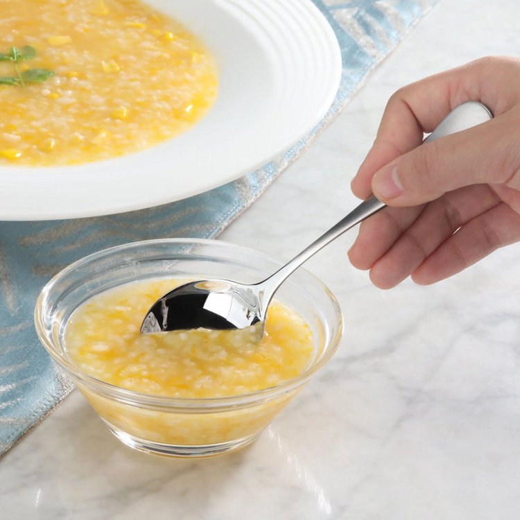Hand holding stainless steel bouillon spoon with matte finish handle, scooping soup from a glass bowl.