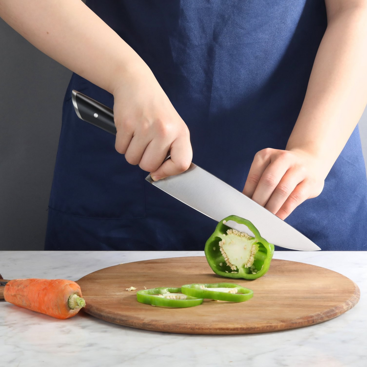 Person wearing a blue apron slicing a green bell pepper on a round wooden cutting board with a large kitchen knife.