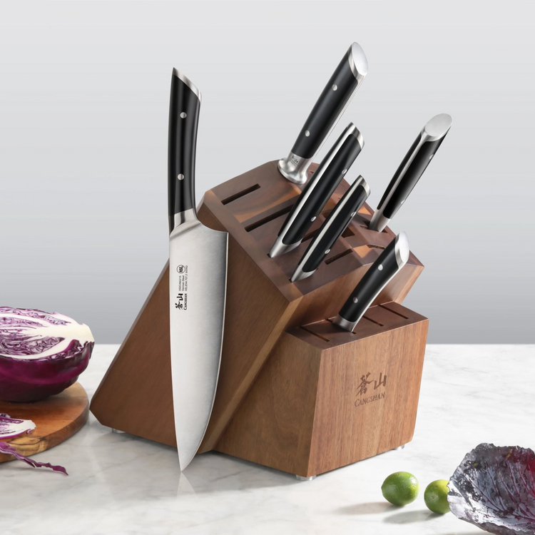 Wooden angled knife block with six knives inserted, placed on a marble surface with cabbage and limes nearby.