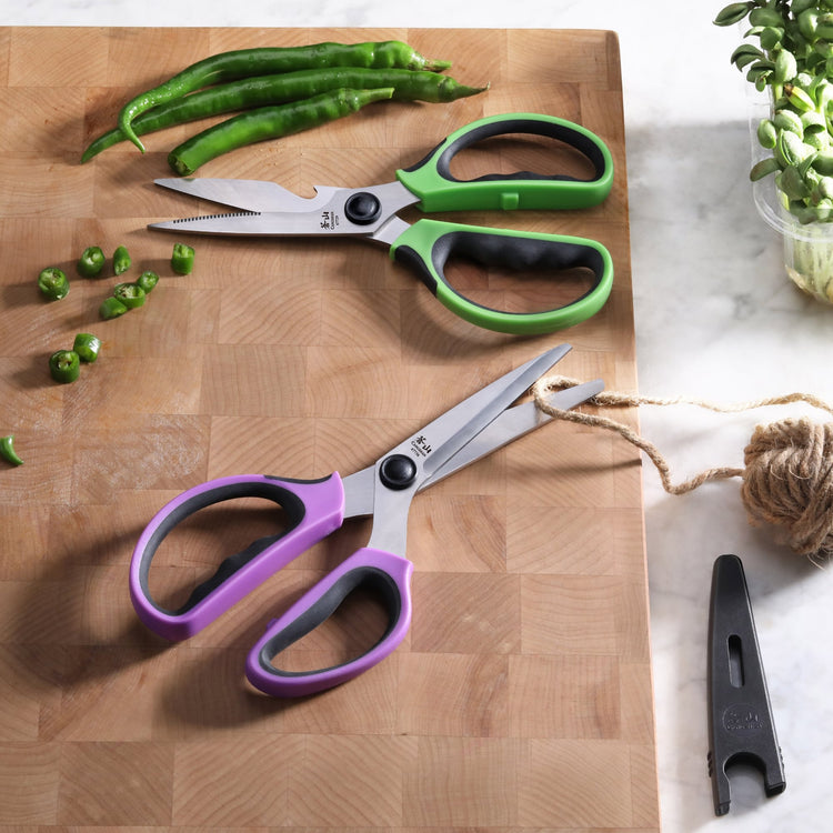 Two pairs of kitchen shears with colorful handles, one green and one purple, on a wooden cutting board with chopped green chi