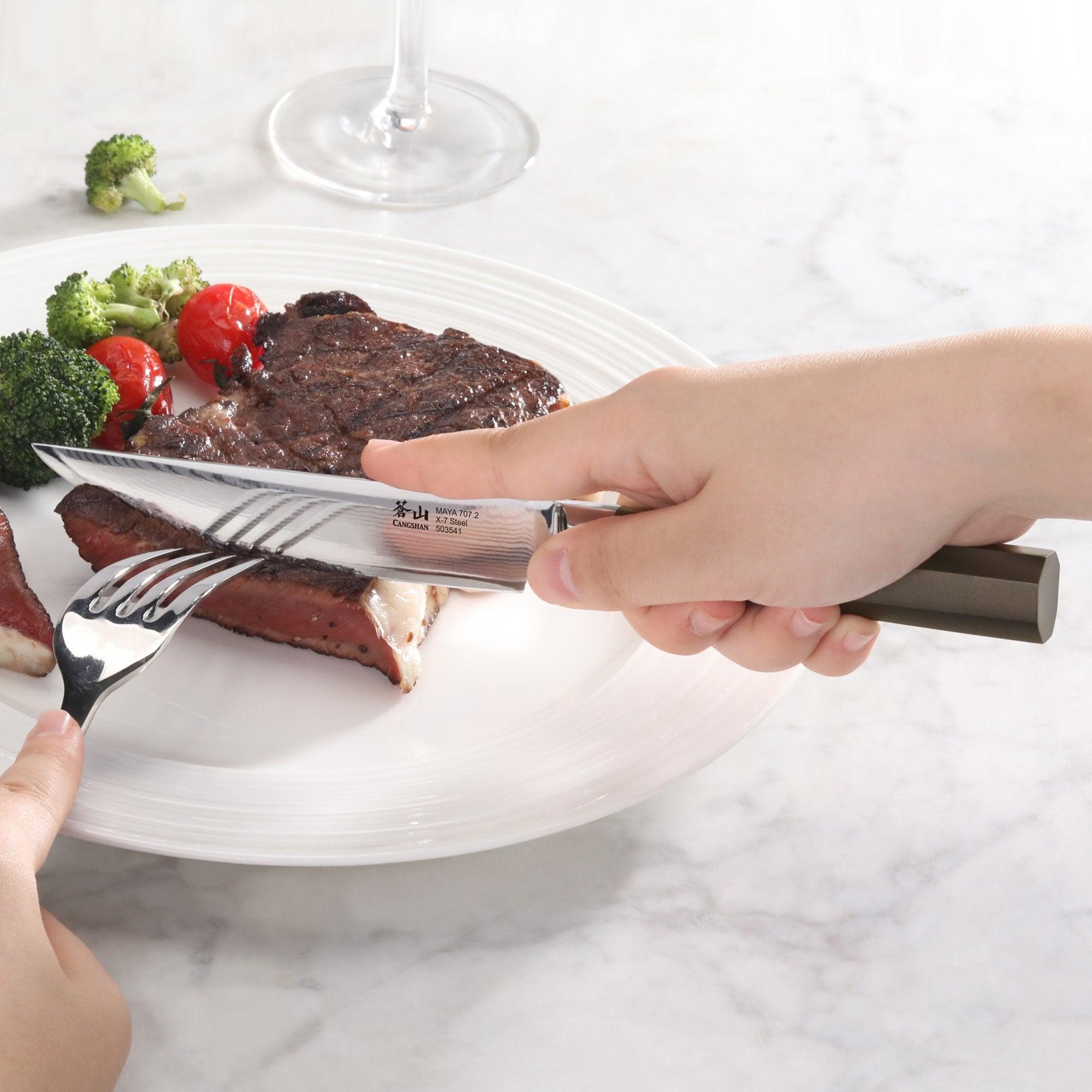 Hand holding a steak knife cutting a medium-rare steak on a white plate with broccoli and cherry tomatoes.
