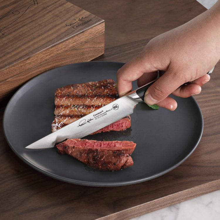 Hand holding a steak knife cutting a medium-rare grilled steak on a black plate next to a wooden storage box.