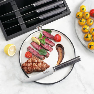 
                    
                      Four steak knives with pakkawood handles in a wooden box, one knife cutting sliced steak on a plate with grilled vegetables.
                    
                  