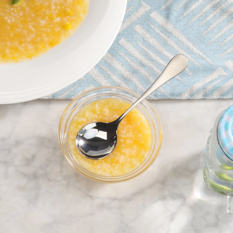 Stainless steel bouillon spoon with matte finish handle resting in a small bowl of soup on a marble surface.